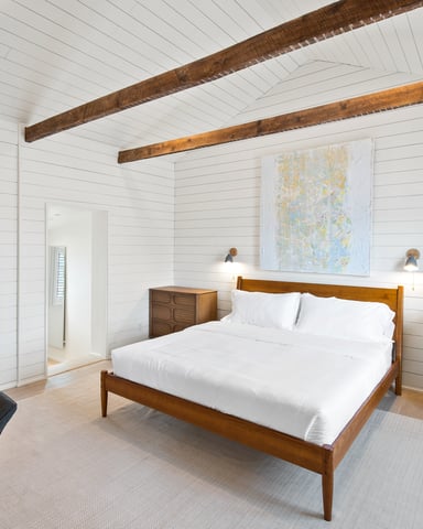 Bedroom with white shiplap walls, exposed wooden beams, wood bed frame, white bedding, and mid-century furniture