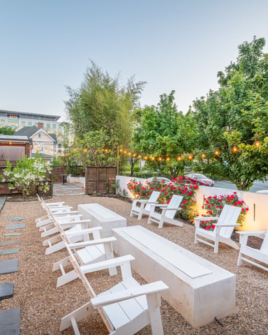 Backyard patio with white Adirondack chairs, concrete benches, red flowering planters, string lights, and brick building at dusk