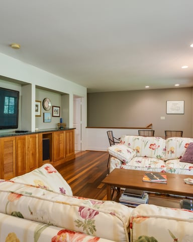 Spacious media room with floral upholstered seating, wooden cabinetry, wall-mounted TV, hardwood floors, and glass doors overlooking trees.