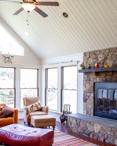 Bright sunroom with vaulted white ceiling, stone fireplace, orange leather sofa, and expansive windows overlooking trees.