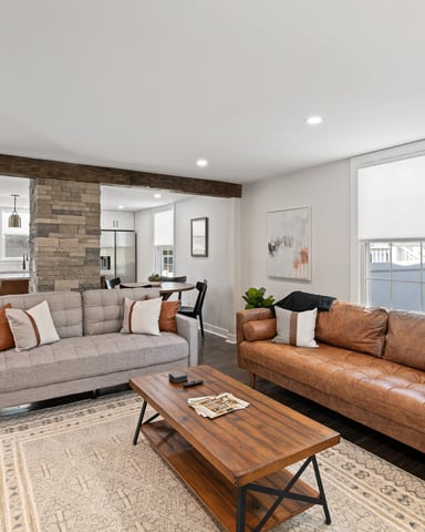 Open-plan living room with gray and cognac leather sofas, wood coffee table, exposed beam, stone fireplace, and kitchen visible in background