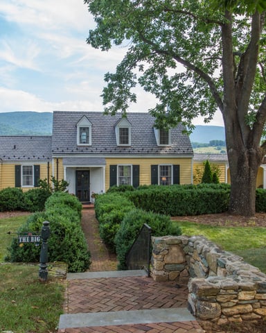 Yellow colonial-style house with gray roof, black shutters, and attached garage, framed by large tree and stone pillars, mountains in background