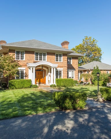 Two-story brick house with white columned entry portico, manicured lawn, and mature trees surrounding property