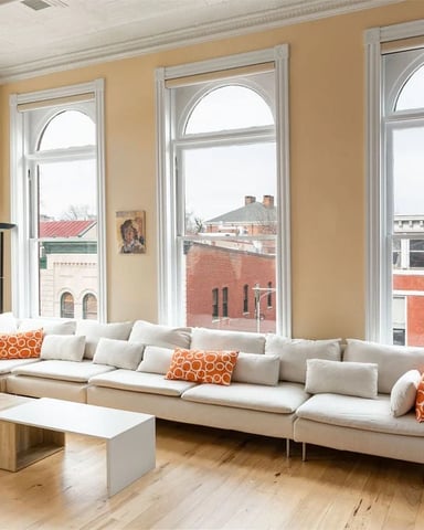 Bright living room with cream sectional sofa, orange accent pillows, wood coffee table, and large windows overlooking brick buildings