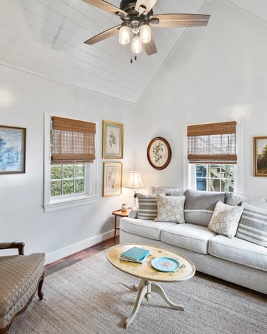 Bright living room with vaulted ceiling, ceiling fan, gray sofa, upholstered armchair, landscape paintings, and windows with woven shades.