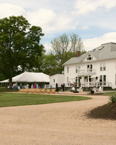 White farmhouse with metal roof and wraparound porches, event tent on lawn, gravel driveway, mature trees and green grounds.