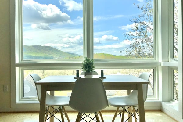 Dining table with three chairs facing large windows overlooking green mountains and blue sky with clouds