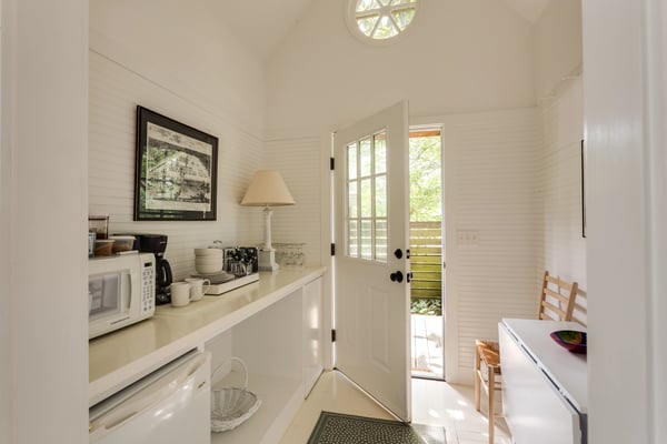 Bright living room with vaulted white ceiling, wood furniture, cream seating, TV on wooden cabinet, and large windows overlooking greenery